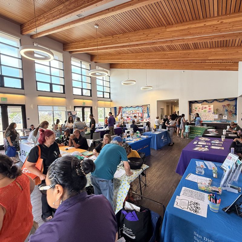 A busy community hall during a resource fair, with neighbors gathered around tables staffed by local service organizations.