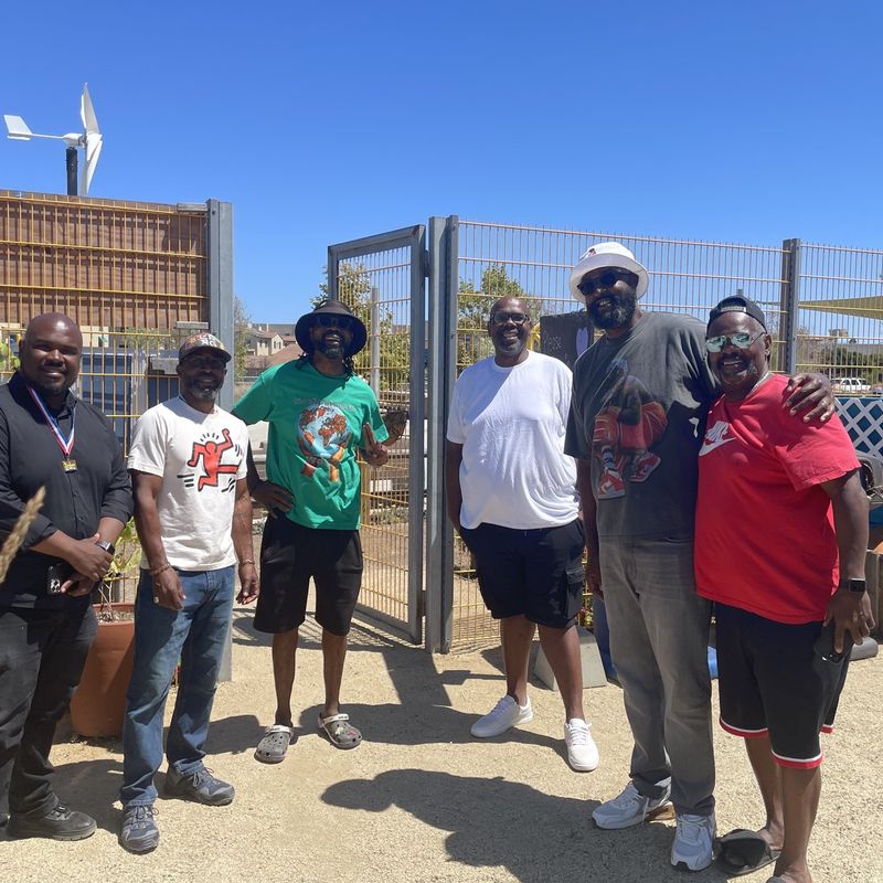 Community members gathered at the open gate of a neighborhood garden on a clear, sunny day.