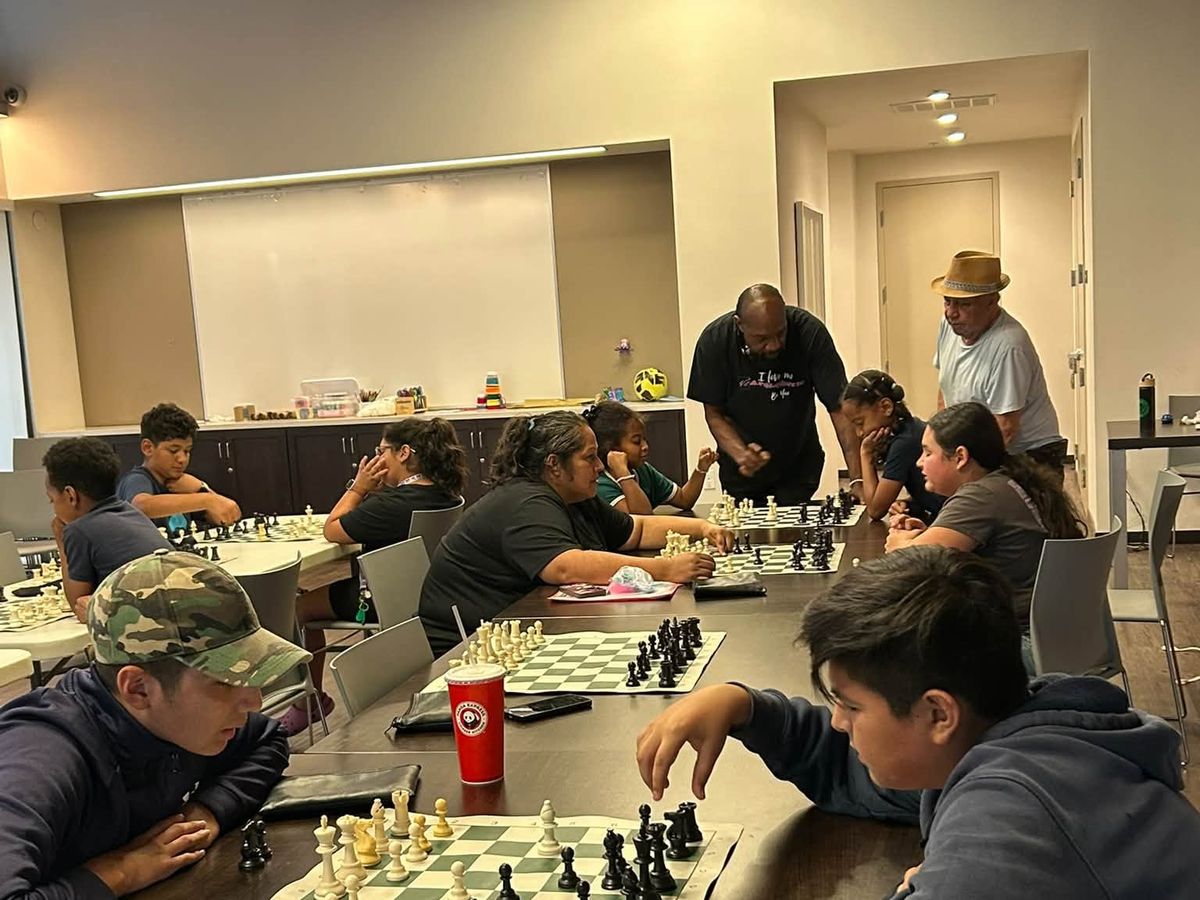 Young players at chess boards while two adult coaches lean in to help during the first Chess at the Creek class.