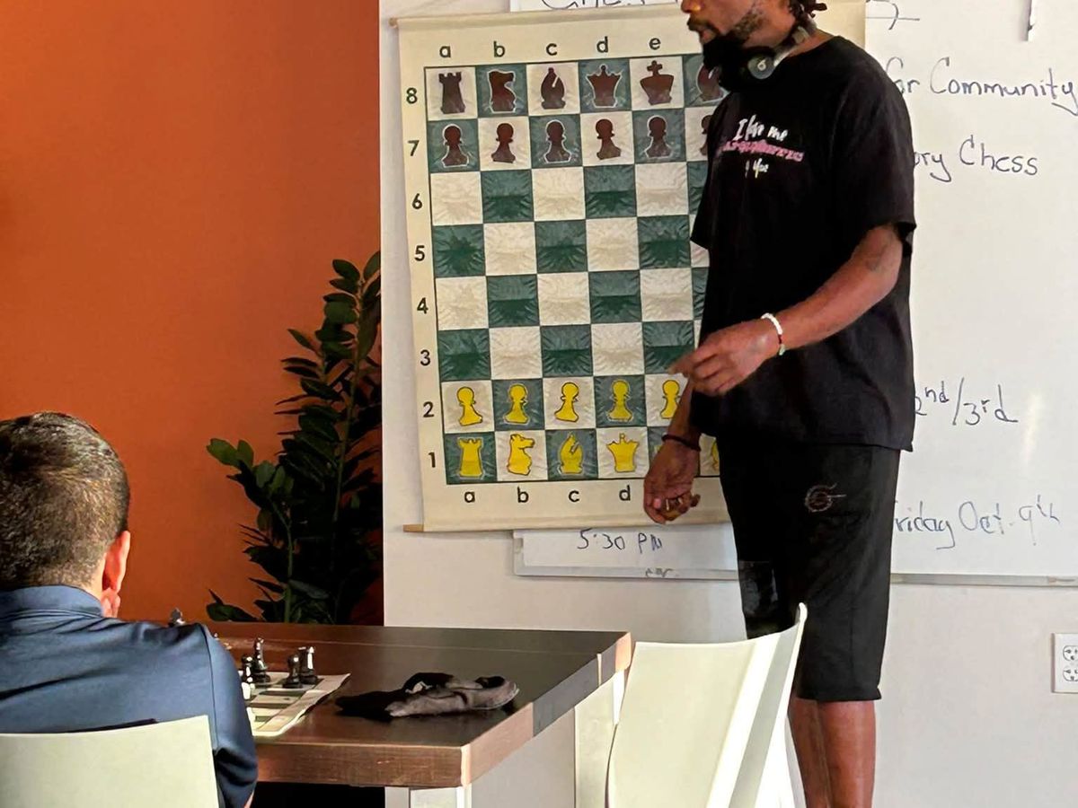 An instructor standing at a wall-mounted demonstration chess board beside a whiteboard during class.