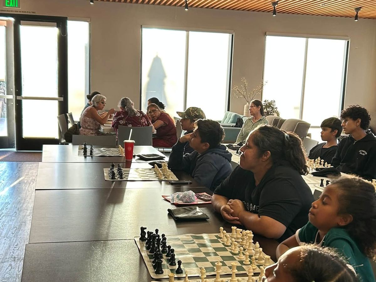 Players and a coach gathered around chess boards at a long table beside tall windows.