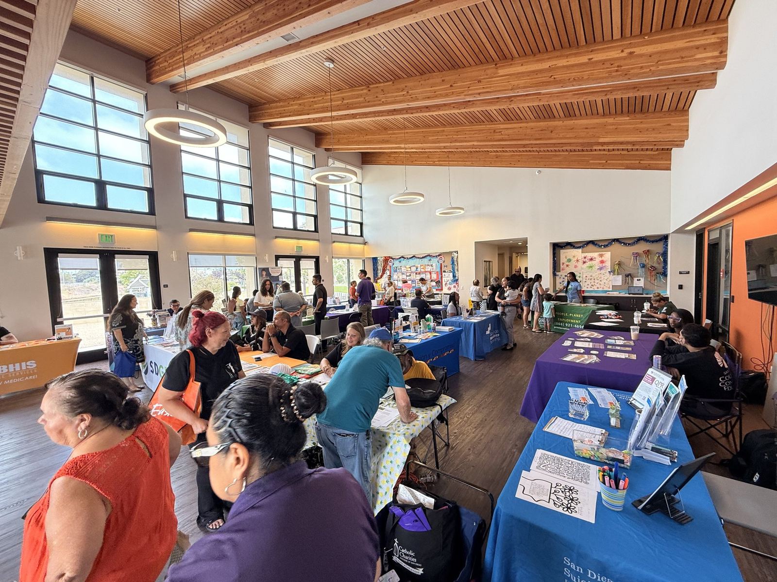 Neighbors and staff talking at information tables inside a community hall during a resource fair.